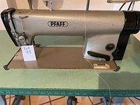 Close-up of PFAFF industrial sewing machine mounted on a table with metal base and green top, showing machine body and thread wire.