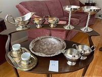 View of silver, copper, and silver plated tableware on wooden shelf with a pink couch background.