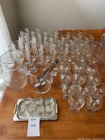 Clear glass drinkware displayed on a wooden surface. Includes multiple champagne flutes featuring an Olympic flame motif, tall water glasses, cognac glasses, a glass pitcher, an ice bucket with tongs, and a small tray holding seven shot glasses.