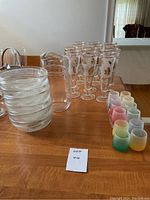 Photo showing six stacked clear ribbed glass bowls, clear glass pitcher, and ten clear etched champagne flutes with gold floral design arranged on a wood table.