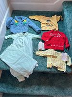 Top-down view of six pieces of vintage baby clothing including cardigans, sweatshirt, sleepsuit, bonnet, and mittens on green carpeted stairs.