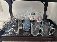 Wide shot showing an assortment of 18 various bar drinking glasses including stemmed, handled mugs, shot glasses, and branded pilsner glasses.