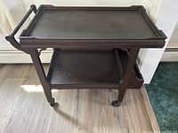 Front view of wooden bar cart with tray top, lower shelf, and wheels on a hardwood floor