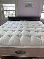 Full view of king size bed with tufted white mattress and wooden headboard between two windows