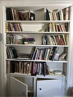 Full view of white bookshelf filled with vintage books ranging from 1950s to 2000s. Books are arranged on multiple shelves with some stacked horizontally. Books show worn condition.