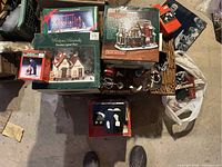 Top-down view showing multiple boxed Christmas village items including the Dickens Keepsake porcelain lighted house and swing set, and other assorted items in a basket on the floor.