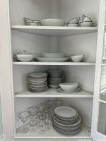 Full view of the dinnerware and glassware set displayed in a white cabinet, showcasing plates, bowls, serving dishes, gravy boat, and glassware.