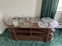 Full view of 14 assorted clear glass serving dishes on a wooden sideboard including cake stands, trays, bowls, and plates of various sizes and shapes.