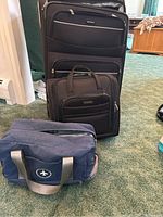 Image showing three travel bags for the lot. A large black upright suitcase with multiple zippered compartments, a smaller black carry-on suitcase also with front pockets, and a small blue duffel bag with handles and airplane logo patch.