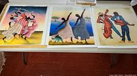 Three signed canvas artworks displayed laid flat on a table, showing colorful abstract dance and music themed images.