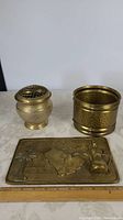 Three brass items on table: wall hanging, flower frog, flower pot holder
