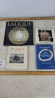 Overview photo of four hardcover collector books laid flat showing front covers: "Lalique" with embossed glass orb, "Fabergé Treasures" jigsaw puzzle book with intricate Fabergé egg image, "The Book of Pearls" in blue, and "Pearls: A First Book" with pearl necklace photo.