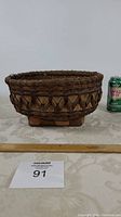 Side view of the handwoven Indigenous basket with a soda can for scale, showing geometric woven pattern and wooden stand base