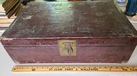 Full frontal view of the dark brown rectangular trunk with visible embossed detailing and brass latch, placed next to a ruler for scale.