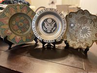 Three vintage plates displayed on stands: steel plate center with U.S. Great Seal, flanked by two large copper plates with floral patterns.