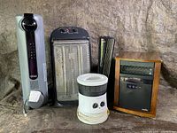 Lot of five assorted electric heaters displayed against a cloth backdrop. Includes tower, panel, fan, cylindrical and cabinet-heater types.
