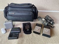 Camera bag with flash units, binoculars and other accessories arranged flat on carpet.