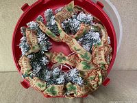 Photo showing the Christmas wreath inside the red storage case from a top angle, displaying the snowy pine branches and beige 'Merry Christmas' ribbon wrapped around it.
