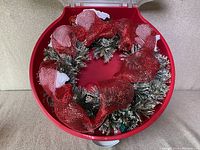 Top view of the wreath inside the red storage case showing the artificial pine branches and decorative red mesh ribbon.