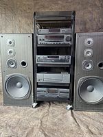 Front view of Kenwood home theater system components stacked in black metal cart, flanked by two large speakers showing speaker cones.