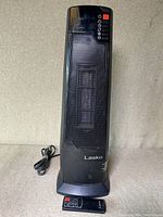 Front view of the Lasko black tower heater with visible control panel and attached power cord, remote control placed in front.