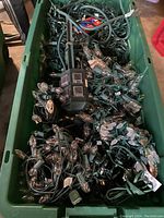 Photo showing a large green plastic container filled with many tangled strings of Christmas lights with green cords and bulbs, along with green extension cords and an over-sized Christmas pin.