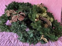Photo showing two green artificial Christmas wreaths with different decorative bows and ornaments, placed on a pink quilted surface.