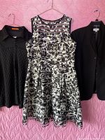 Three women's clothing items hanging on hangers against a pink background: black pleated blouse, black and white floral sleeveless dress, black blazer jacket.