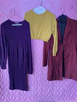 Purple dress, mustard cropped sweater, and long red blazer hung on hangers against pink quilted background.