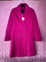 Full view of hot pink wool coat hanging on a hanger, showing the front with buttons and pockets.