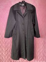 Front view of black women's wool coat laid flat on a pink quilted surface, showing the full length, collar, buttons, and side pockets.