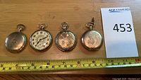 Four gold-tone pocket watches laid side by side with measuring tape and lot card