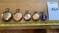 Five pocket watches arranged in a row on wooden surface with measuring tape.