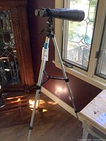 Full view of black binoculars mounted on silver tripod stand in front of window, label showing lot number attached to tripod.