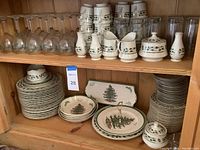 Photo showing multiple pieces of Christmas-themed china including plates, bowls, creamers, sugar bowls, small pitchers, clear tumblers, and wine glasses organized on wooden shelves.