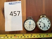 Two pocket watches side by side on wooden surface with measuring tape showing dimensions. Left: Ford V8 logo silver‐tone watch; right: Hamilton gold‐tone watch.