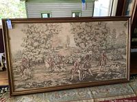 Wide shot of the entire framed needlepoint tapestry showing hunting figures, horses, and dogs in a natural wooded landscape
