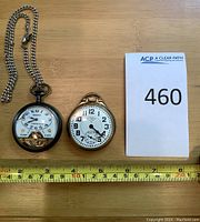 Two pocket watches on wooden surface with measuring tape and lot card visible
