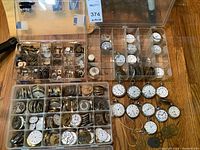 Overhead view of two clear plastic organizers with multiple pocket watch movements and loose watches and parts on wooden surface