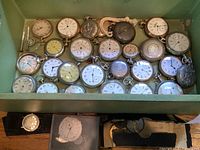 Green tray holding approximately two dozen pocket watch cases and dials