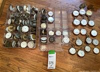 Overhead view of multiple plastic trays filled with pocket watch movements, dials and cases arranged on wood floor alongside complete watches.