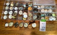 Two clear plastic boxes on wooden table with multiple compartments filled with pocket watch movements, dials and cases; row of eight assembled pocket watches in front with lot tag