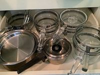 Drawer with clear glass cookware: four heatproof glass pots with locking handles and matching lids, plus two stainless steel pans with black handles and lids