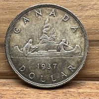 Reverse side of 1937 Canadian silver dollar showing canoe with two paddlers, bundles, and trees with inscription 'CANADA DOLLAR 1937'.