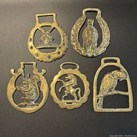 Five brass horse brasses laid out together showing the designs including windmill, man figure, squirrel, horse, and bird.