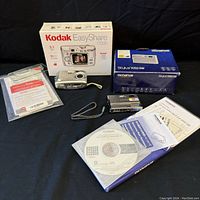 Kodak EasyShare C7330 camera, box, manual, and Olympus Stylus 1050 SW box with accessories on black background.