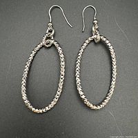 Pair of sterling silver drop earrings shown from front on black background highlighting textured oval hoops and hook ear wires.