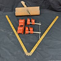 Full view of all three items: the folded wooden ruler with brass joints, the red pipe clamps arranged side by side, and the vintage wooden moulding plane with handle and blade.