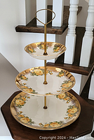 Front view of three-tier cake stand showing all three plates with yellow rose floral designs and gold accents mounted on gold metal frame