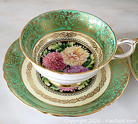 Side view of cup resting on saucer showing green enamel border, gold gilt filigree, and floral center motif